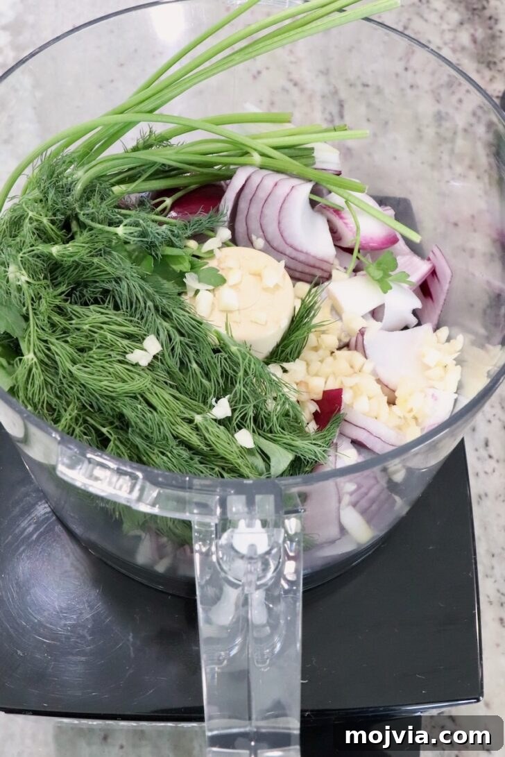 Greek Gyro Meatballs Recipe — Flavorful Mediterranean Meatballs 6 A food processor bowl filled with fresh herbs, sliced red onions, garlic, and parsley on a kitchen counter.