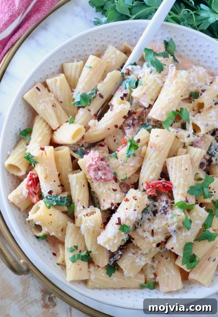 Creamy Tuscan Chicken Pasta Recipe with Sun-Dried Tomatoes 2 A bowl of creamy rigatoni pasta topped with fresh parsley, grated cheese, and red pepper flakes, mixed with sun-dried tomatoes; a fork rests in the bowl.