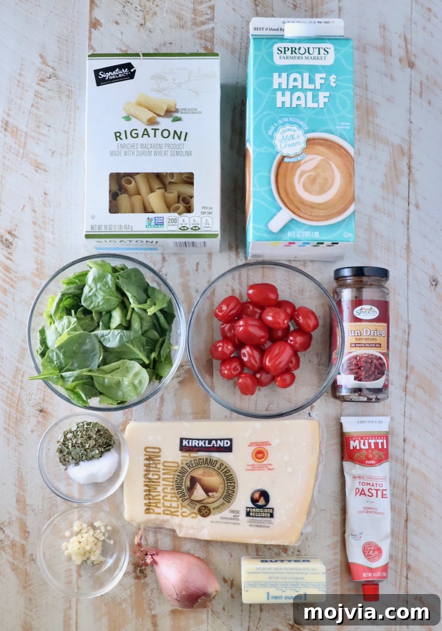 Creamy Tuscan Chicken Pasta Recipe with Sun-Dried Tomatoes 5 Assorted ingredients on a light wooden surface, including rigatoni, half & half, spinach, cherry tomatoes, garlic, herbs, parmesan, tomato paste, shallot, butter, and sun-dried tomatoes.
