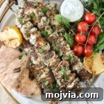 Authentic Greek Souvlaki Recipe: How to Make Classic Meat Skewers 11 grilled chicken souvlaki on a plate with pita bread, a small bowl of tzatziki sauce, tomatoes, lemons, and herbs