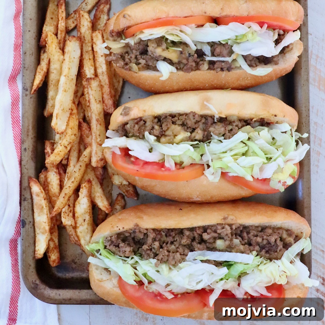 three chopped cheese sandwiches with sliced tomatoes and shredded lettuce lined up on a baking sheet
