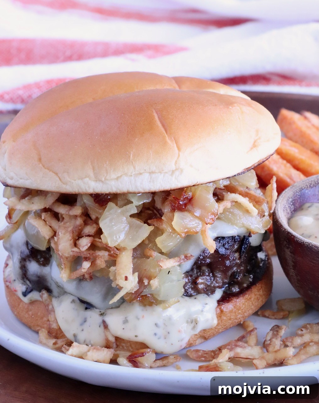 French Onion Burger Recipe with Caramelized Onions & Gruyère 2 cheeseburger on a plate topped with caramelized onions and crispy fried onions, with a small bowl of sauce on the side