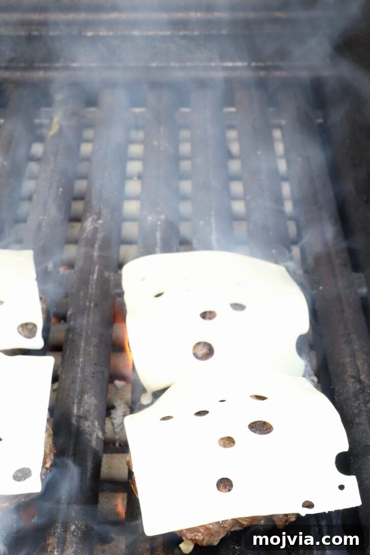 French Onion Burger Recipe with Caramelized Onions & Gruyère 9 burger patties on a grill topped with sliced swiss cheese