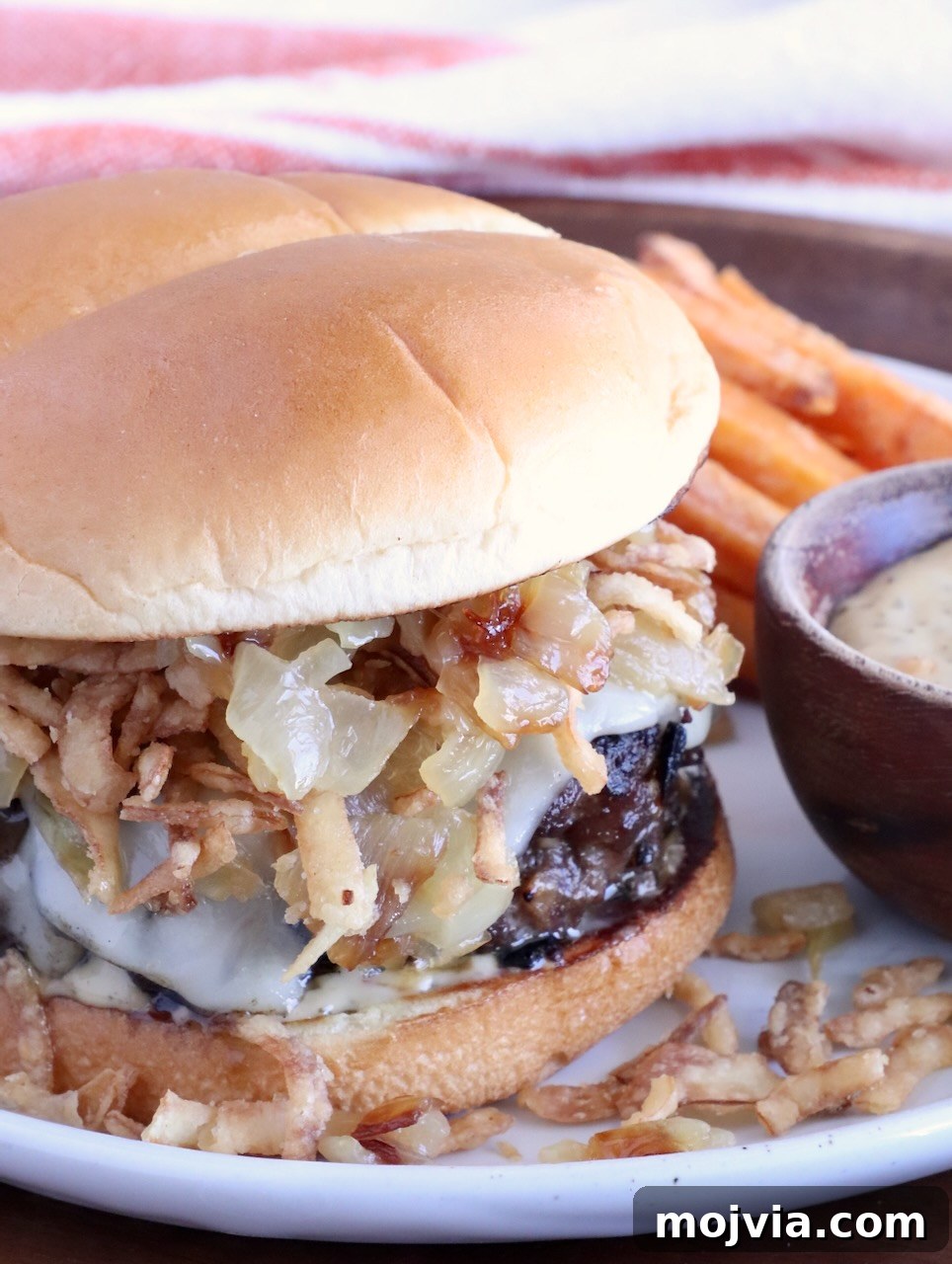 French Onion Burger Recipe with Caramelized Onions & Gruyère 10 cheeseburger on a plate topped with crispy onions, with a side bowl of sauce, and sweet potato fries behind the burger on the plate