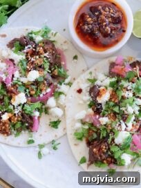 Instant Pot Beef Barbacoa: Tender Mexican Shredded Beef 13 two tacos on a plate topped with beef barbacoa, crumbled cheese, pickled onions, salsa macha, and cilantro