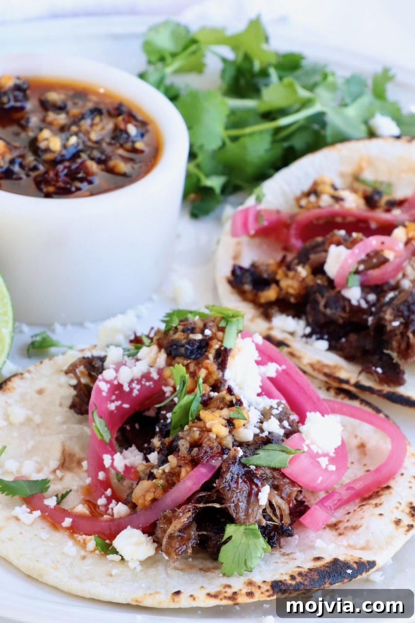 Tender Smoked Beef Cheek Barbacoa Recipe 2 two tacos on a plate topped with smoked beef cheek barbacoa, pickled onions, salsa, cotija cheese and cilantro