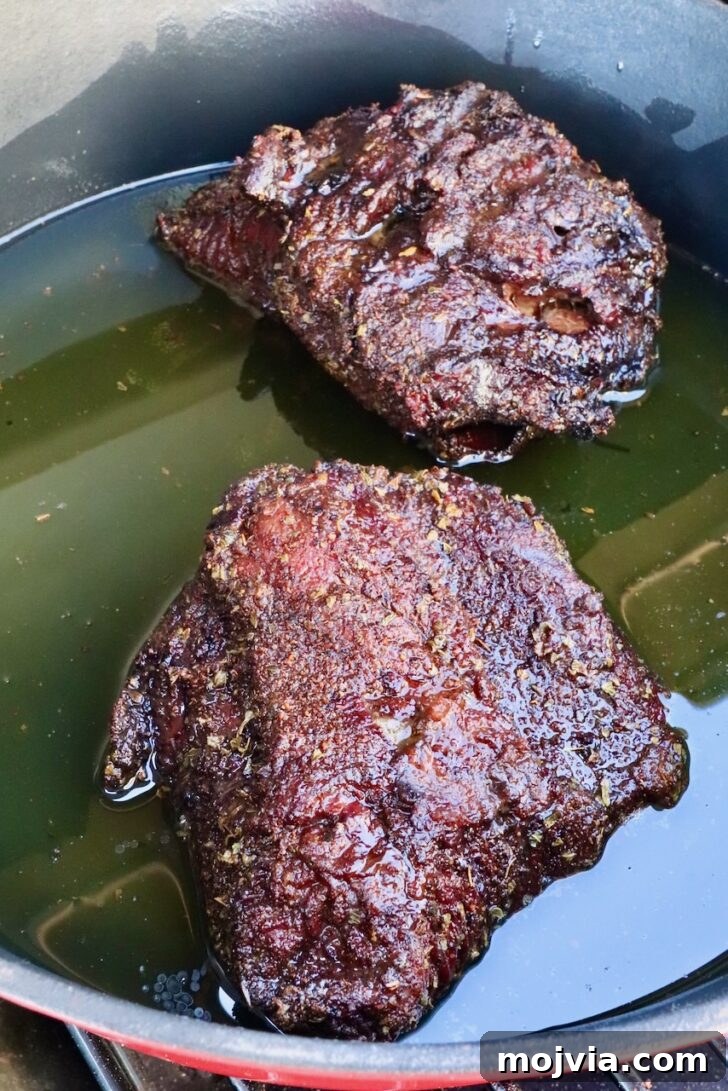 Tender Smoked Beef Cheek Barbacoa Recipe 8 beef cheeks sitting in melted beef tallow in a cast iron skillet on a smoker