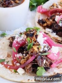 Tender Smoked Beef Cheek Barbacoa Recipe 10 smoked beef cheek tacos on a plate topped with crumbled cheese, fresh cilantro, pickled onions, and salsa